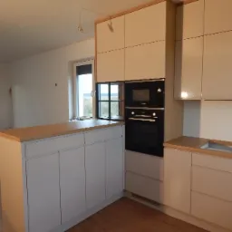 Jasna kuchnia z drewnianym blatem, fronty szafek w odcieniach bieli, zabudowany piekarnik marki Bosch, widok na okno i niedokończone ściany w tle.