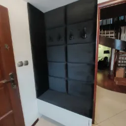 Wnęka z tapicerowanym panelem ściennym w kolorze grafitowym, z siedziskiem i wieszakami, umieszczona przy wejściu do domu, widoczna obok brązowych drzwi wejściowych i schodów na piętro.