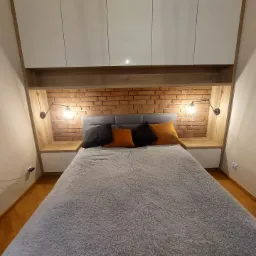 Sypialnia na poddaszu z łóżkiem, ceglaną ścianą, szafkami i lampkami ściennymi w stylu loft, oświetlona ciepłym światłem.