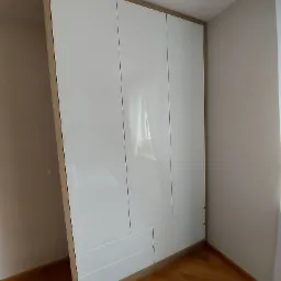 Biała, trzydrzwiowa szafa z połyskiem, z drewnianą ramą, umieszczona w rogu pokoju z parkietem na podłodze. W drzwiach szafy odbija się okno z zasłoną.