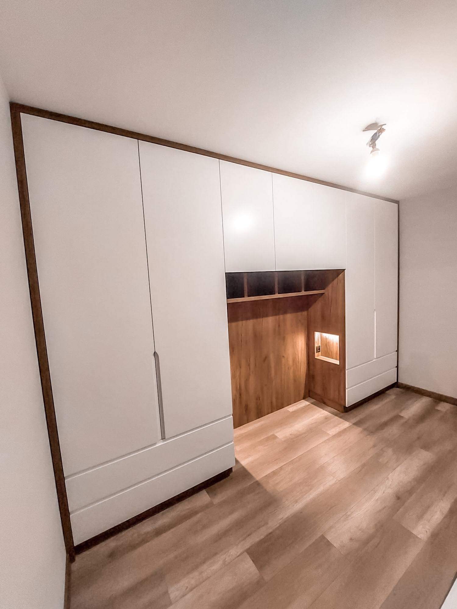 Biała szafa wnękowa z drewnianym obramowaniem i wnęką z podświetleniem, w minimalistycznym wnętrzu z drewnianą podłogą.