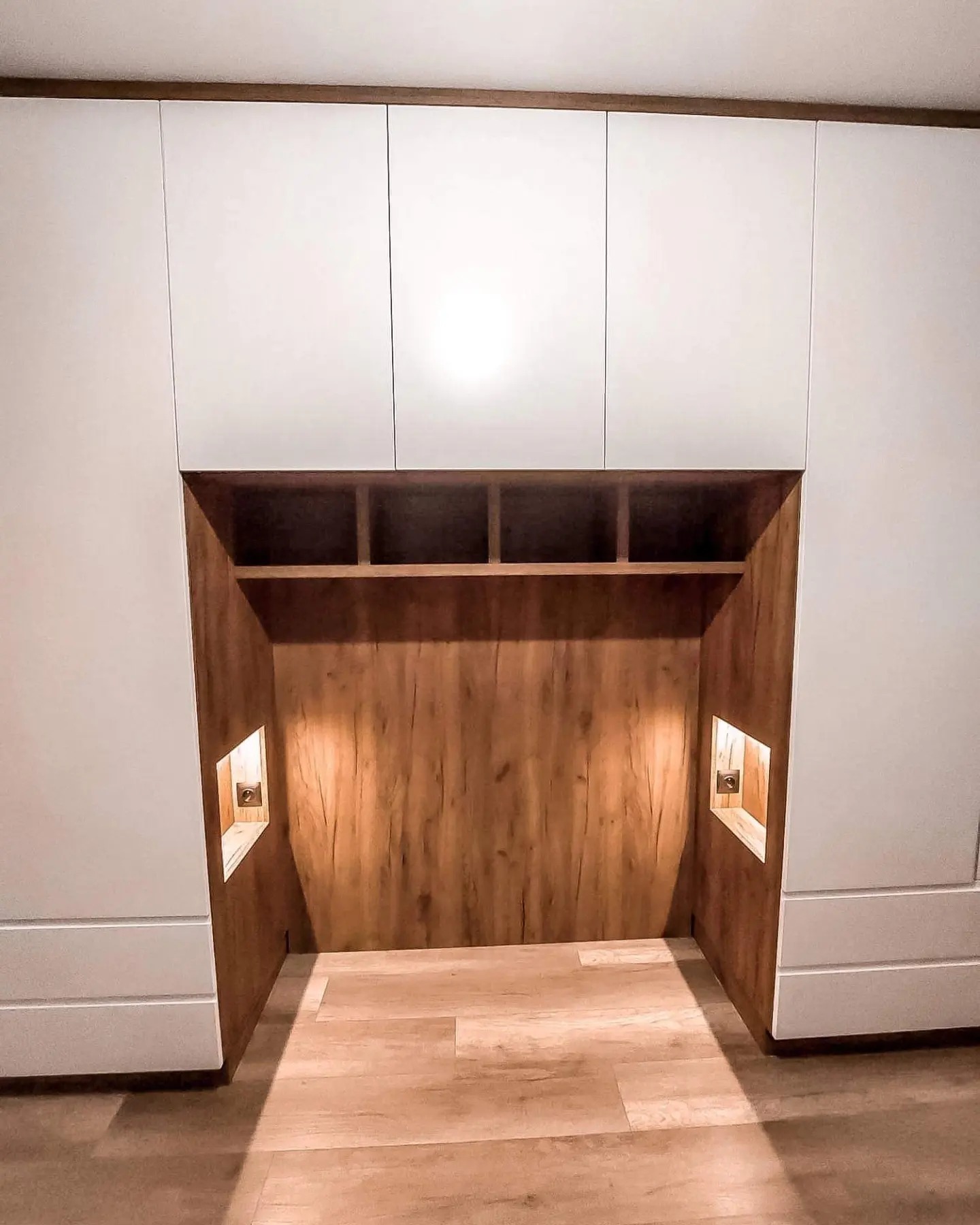 Biała zabudowa meblowa z wnęką wykończoną drewnem, oświetlenie punktowe wewnątrz wnęki, widoczne półki i gniazdko elektryczne.