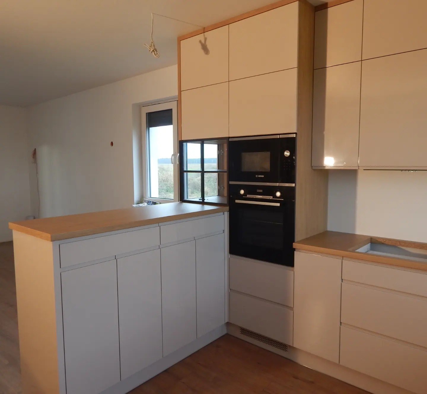 Jasna kuchnia z drewnianym blatem, fronty szafek w odcieniach bieli, zabudowany piekarnik marki Bosch, widok na okno i niedokończone ściany w tle.