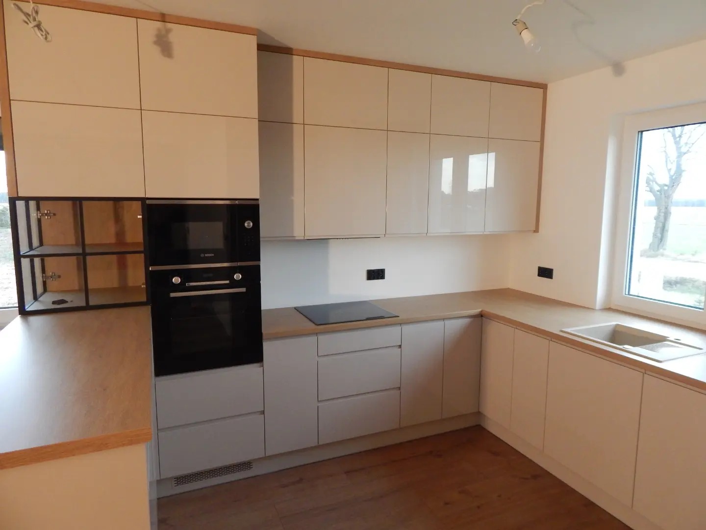 Jasna kuchnia na wymiar z drewnianymi akcentami, widoczne szafki wiszące i stojące, zabudowany piekarnik Bosch, płyta indukcyjna i otwarte półki z czarną ramą.