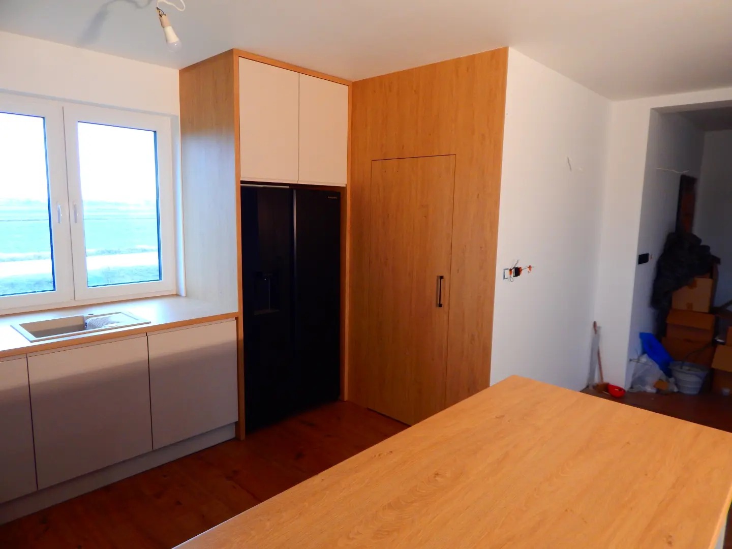 Jasna kuchnia z drewnianymi elementami, białe szafki wiszące nad czarną lodówką, widok na okno i drewniany blat stołu w pierwszym planie.