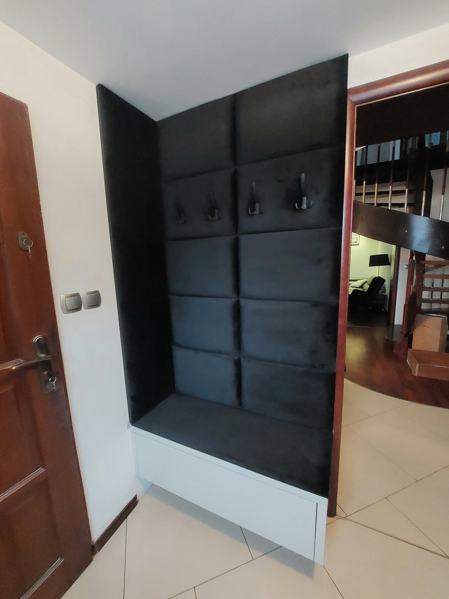 Wnęka z tapicerowanym panelem ściennym w kolorze grafitowym, z siedziskiem i wieszakami, umieszczona przy wejściu do domu, widoczna obok brązowych drzwi wejściowych i schodów na piętro.