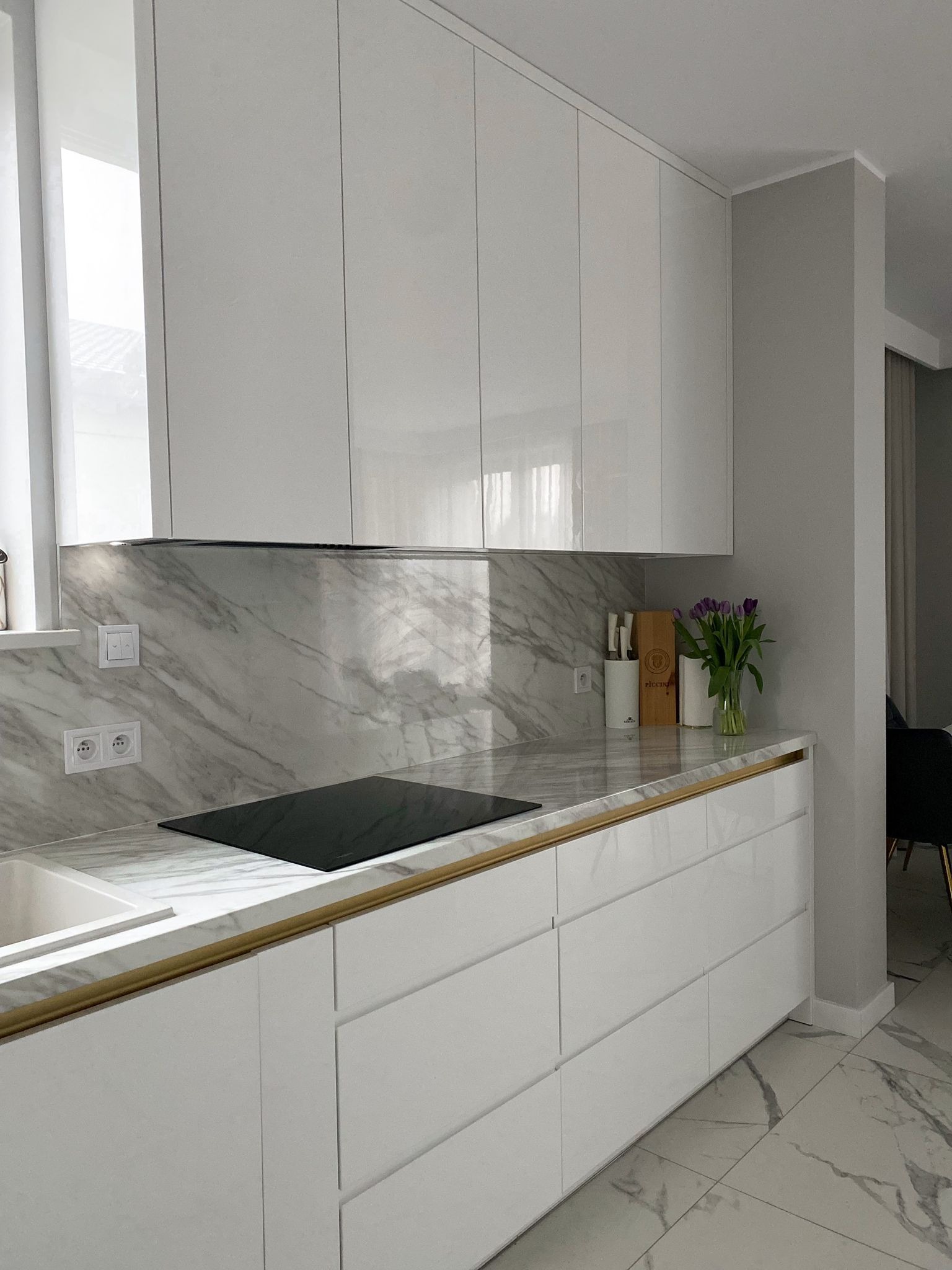 Biała kuchnia z połyskiem, marmurowy blat i ściana nad blatem, złote akcenty, indukcja, kwiaty w wazonie.
