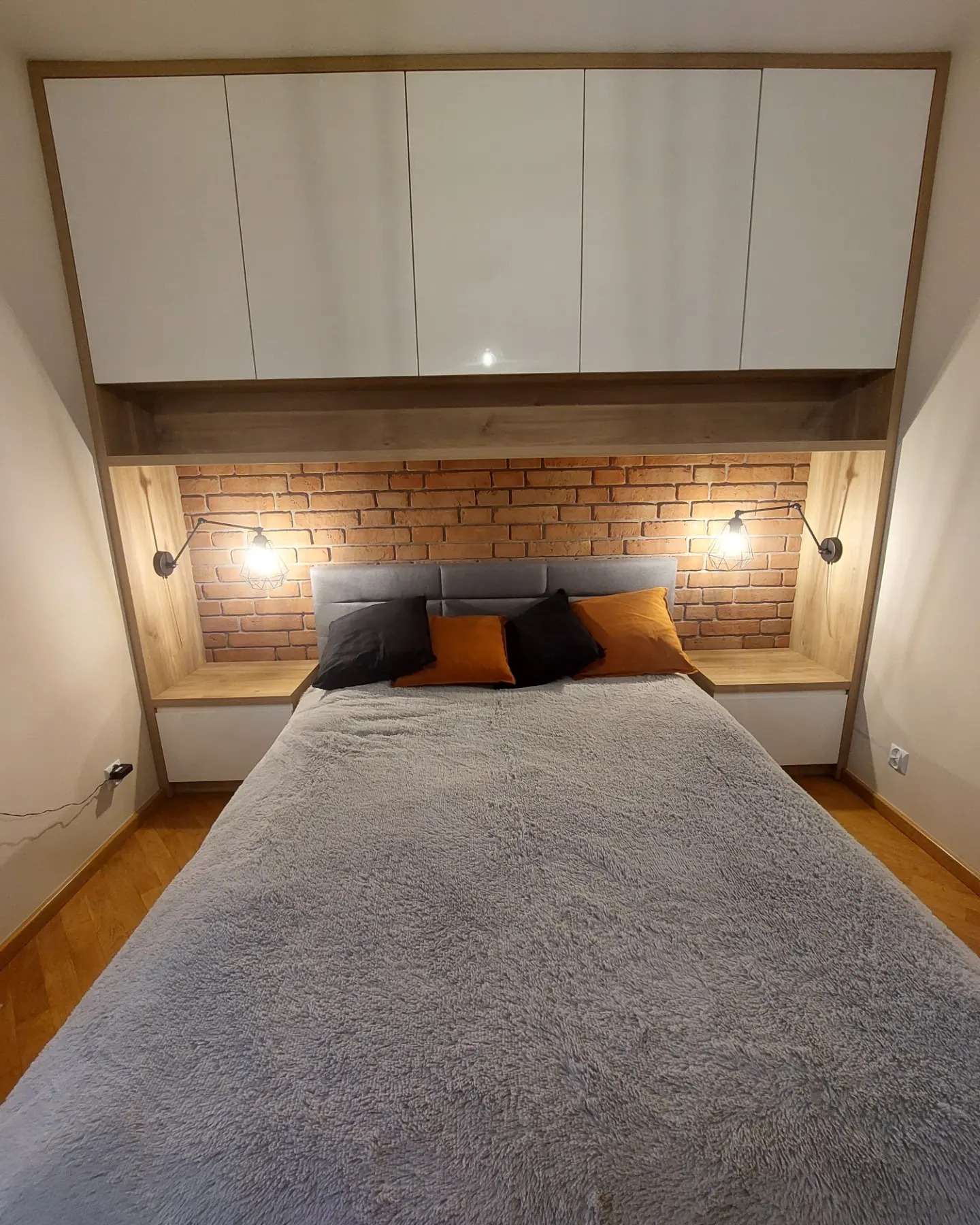 Sypialnia na poddaszu z łóżkiem, ceglaną ścianą, szafkami i lampkami ściennymi w stylu loft, oświetlona ciepłym światłem.