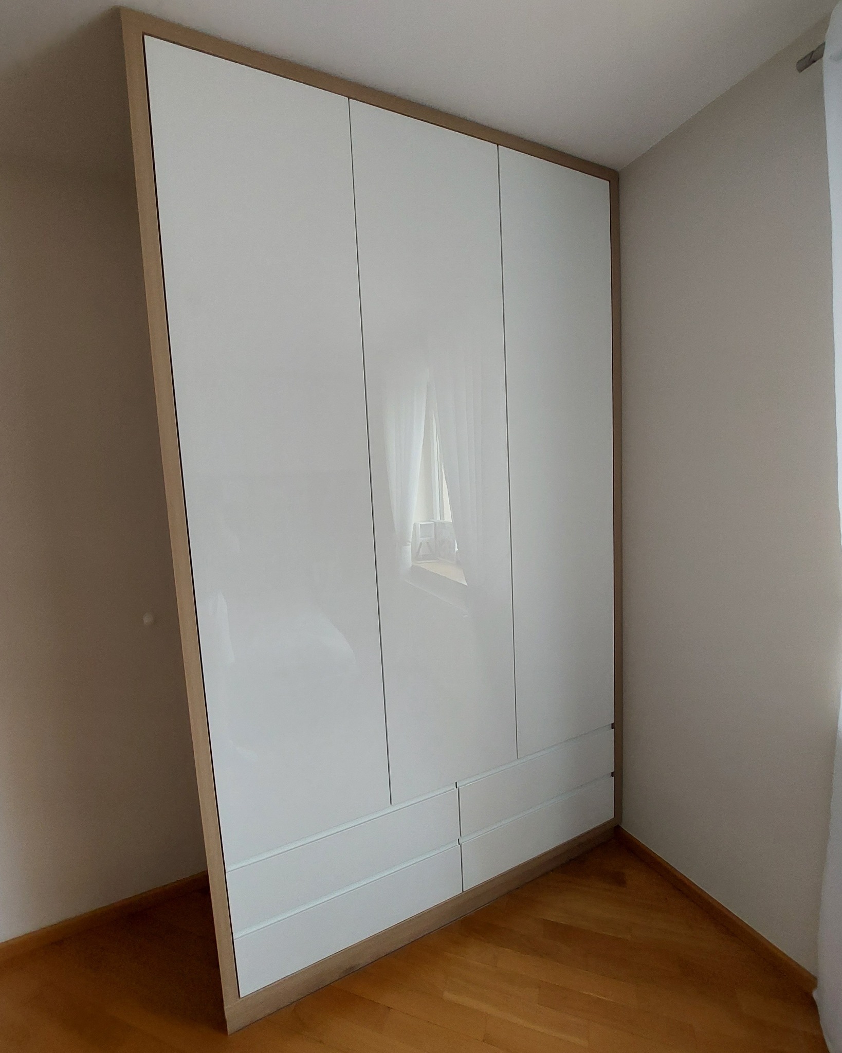 Biała, trzydrzwiowa szafa z połyskiem, z drewnianą ramą, umieszczona w rogu pokoju z parkietem na podłodze. W drzwiach szafy odbija się okno z zasłoną.