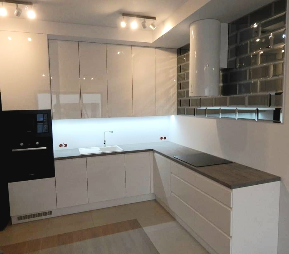 Nowoczesna kuchnia z białymi lakierowanymi frontami, szarym blatem i czarnym piekarnikiem w zabudowie, oświetlona listwą LED pod szafkami wiszącymi, z okapem cylindrycznym na tle szarej ściany...