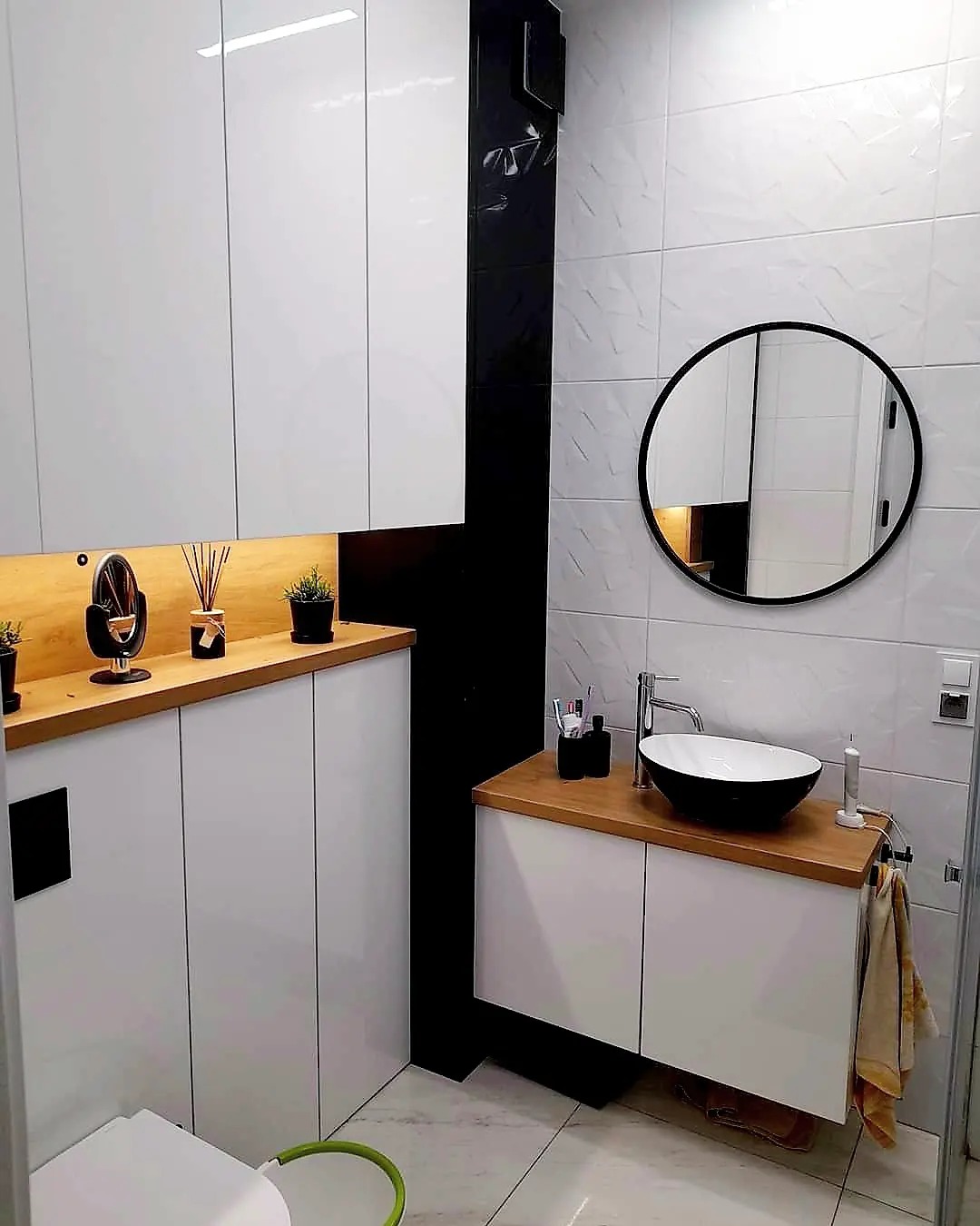Nowoczesna łazienka z białymi szafkami, drewnianym blatem, okrągłym lustrem i czarnymi akcentami, w tym kolumna i umywalka.