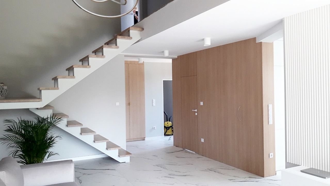 Nowoczesne wnętrze domu z drewnianymi schodami wspornikowymi, jasną podłogą imitującą marmur, i zabudową z jasnego drewna z ukrytymi drzwiami.
