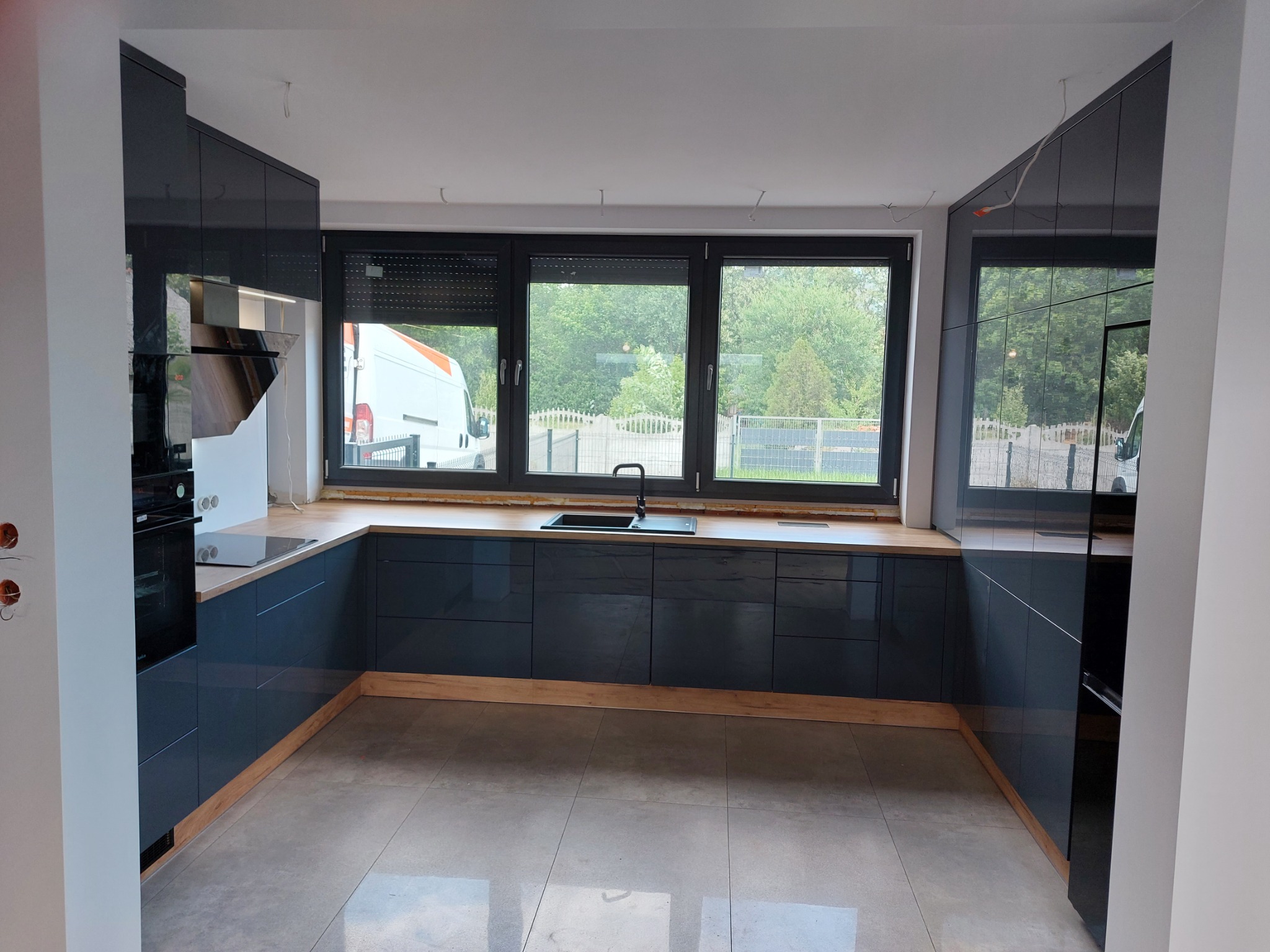Nowoczesna kuchnia na wymiar z ciemnymi, lakierowanymi frontami, drewnianym blatem i widokiem na zewnątrz przez okno, w tle częściowo widoczny biały samochód dostawczy.