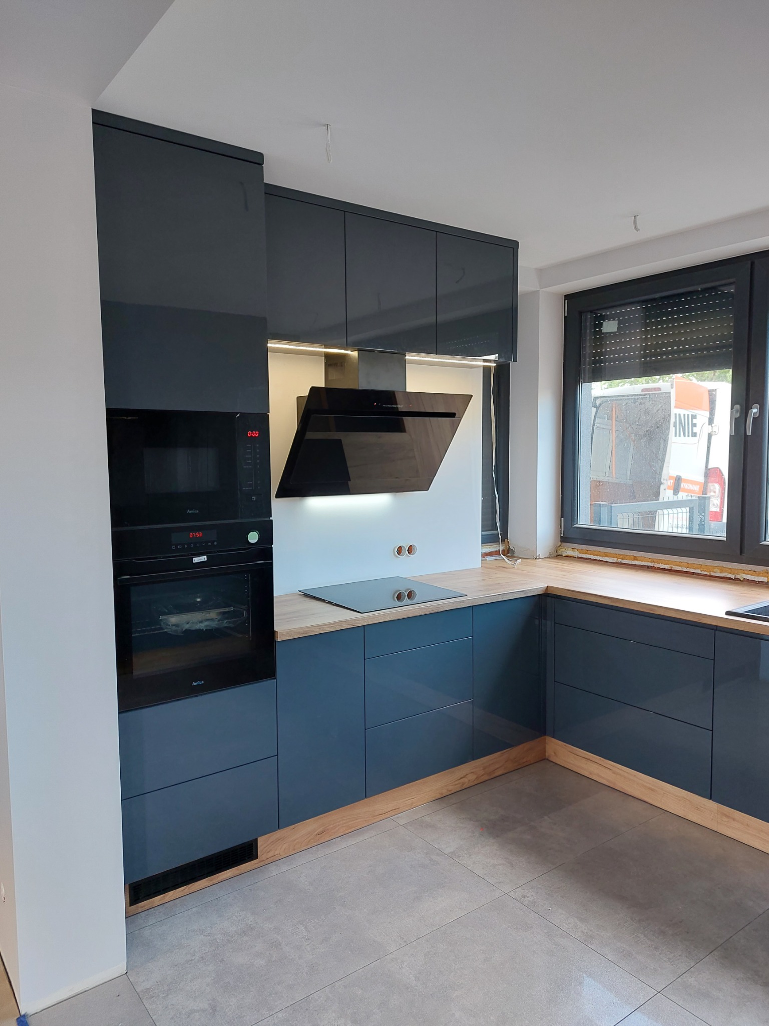 Nowoczesna kuchnia z ciemnogranatowymi, lakierowanymi frontami, drewnianym blatem i czarnym okapem, widok na zabudowę z piekarnikiem i mikrofalówką marki Amica, okno z roletami i samochód dostawczy...