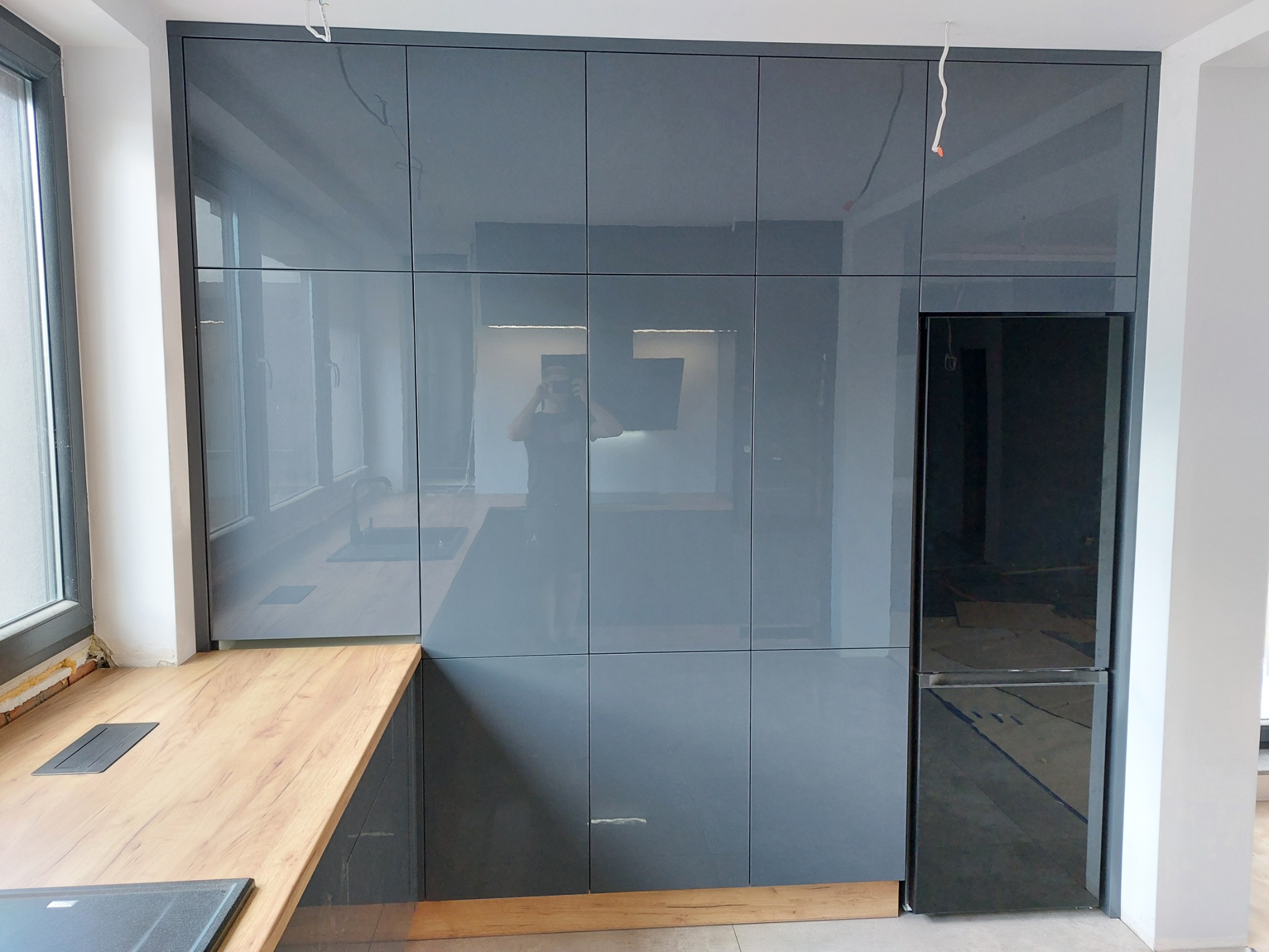 Szare, lakierowane fronty zabudowy kuchennej z wbudowaną lodówką, drewniany blat, widoczne odbicie osoby robiącej zdjęcie.
