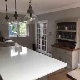 Jasna kuchnia z białym blatem wyspy, trzema wiszącymi lampami z przezroczystego szkła i drewnianymi elementami, oraz kredensem w stylu vintage z przeszklonymi drzwiami w tle.