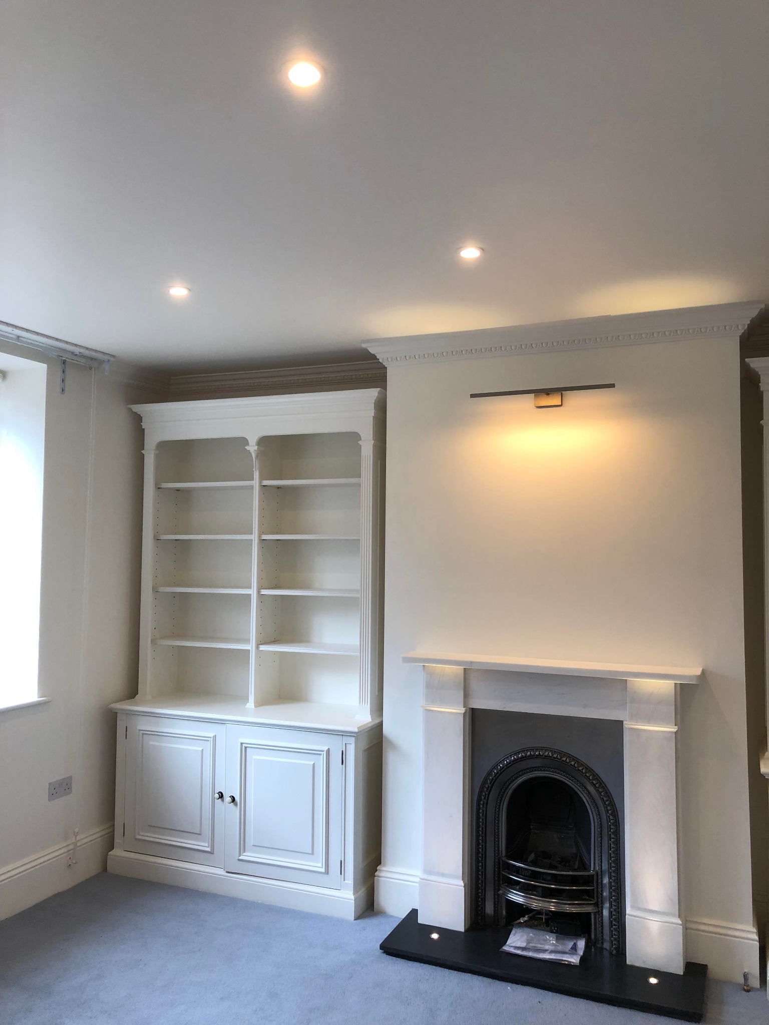 Biała, wbudowana biblioteczka z półkami i szafką na dole, obok marmurowego kominka z czarnym wkładem, oświetlona punktowymi lampami sufitowymi i lampą nad kominkiem w pokoju po odświeżeniu.