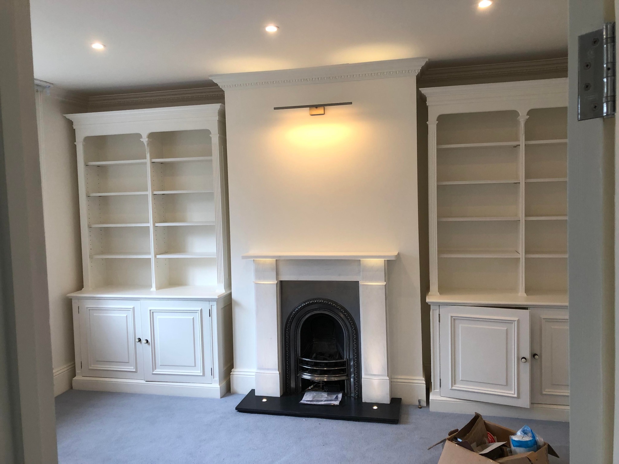 Salon z zabudową biblioteczną po obu stronach kominka z czarnym wkładem, wykończony w bieli, z delikatnym oświetleniem punktowym i listwą nad kominkiem. Jedne z drzwiczek szafki są niedomknięte.