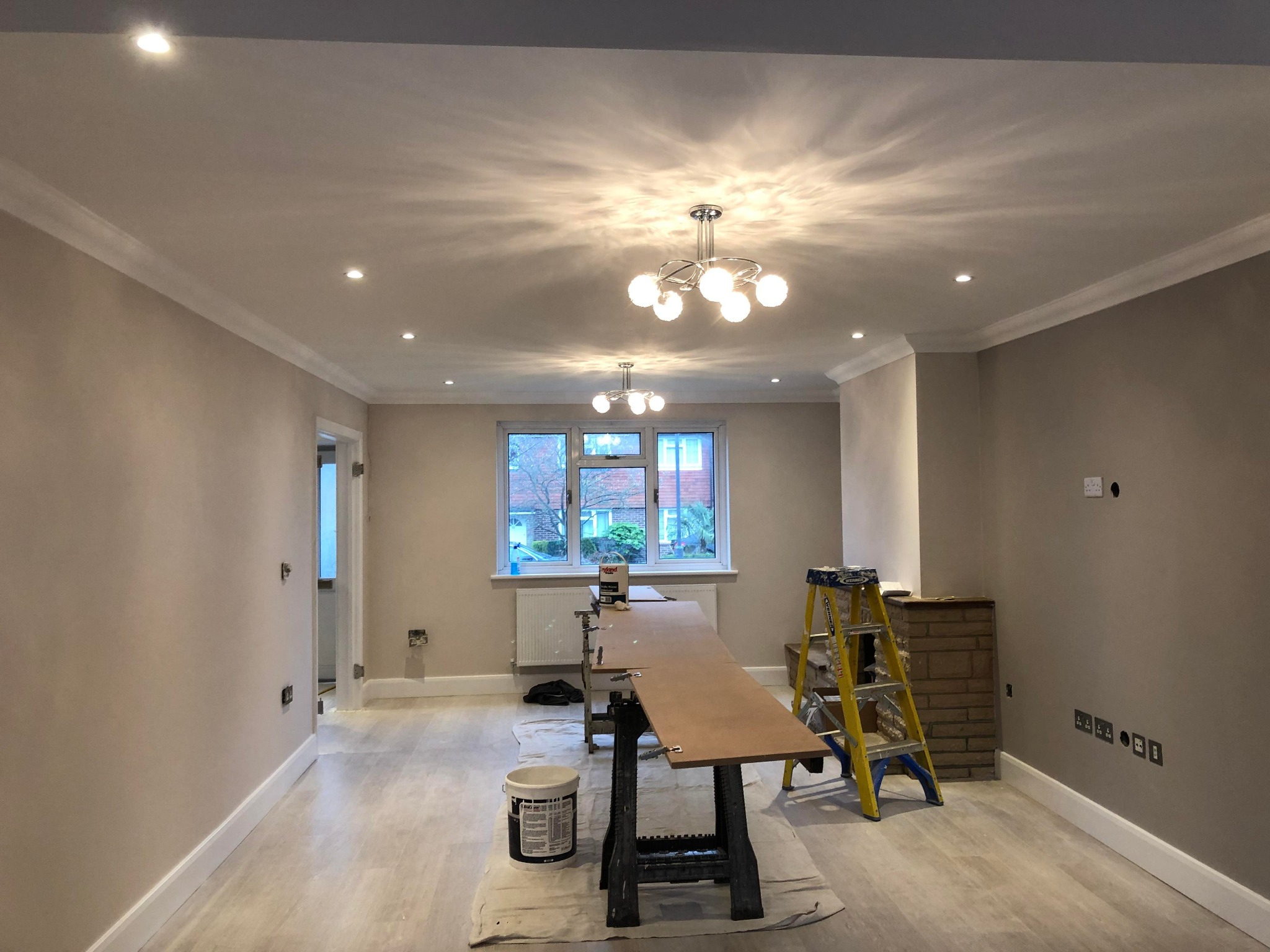 Pokój w trakcie malowania ścian na beżowo, widoczne narzędzia malarskie, drabina, lampy sufitowe i okno z widokiem na zewnątrz.