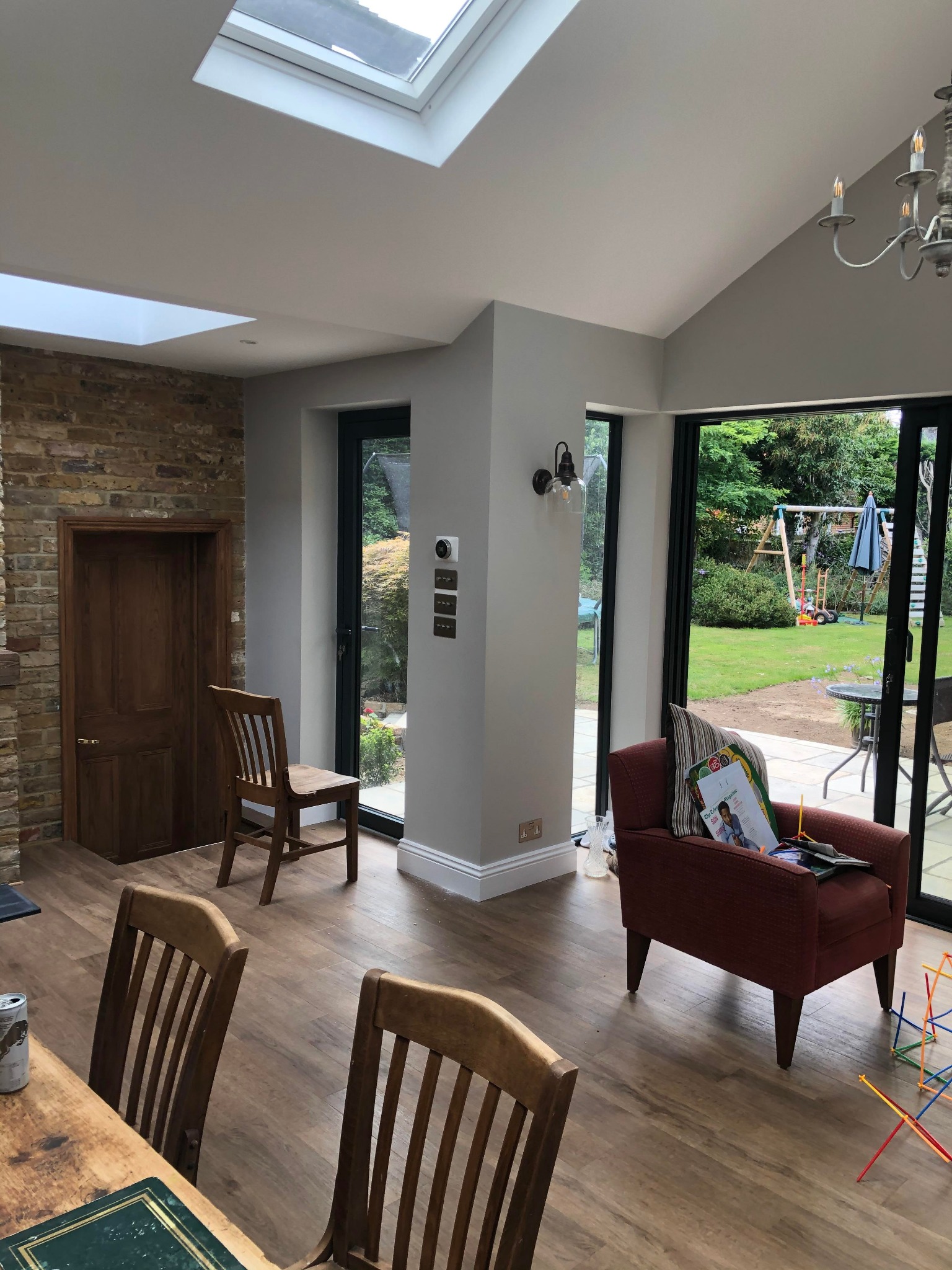 Jasne, przestronne wnętrze po renowacji z drewnianą podłogą, ceglaną ścianą, dużymi oknami z widokiem na ogród z placem zabaw, i nowoczesnym oświetleniem.