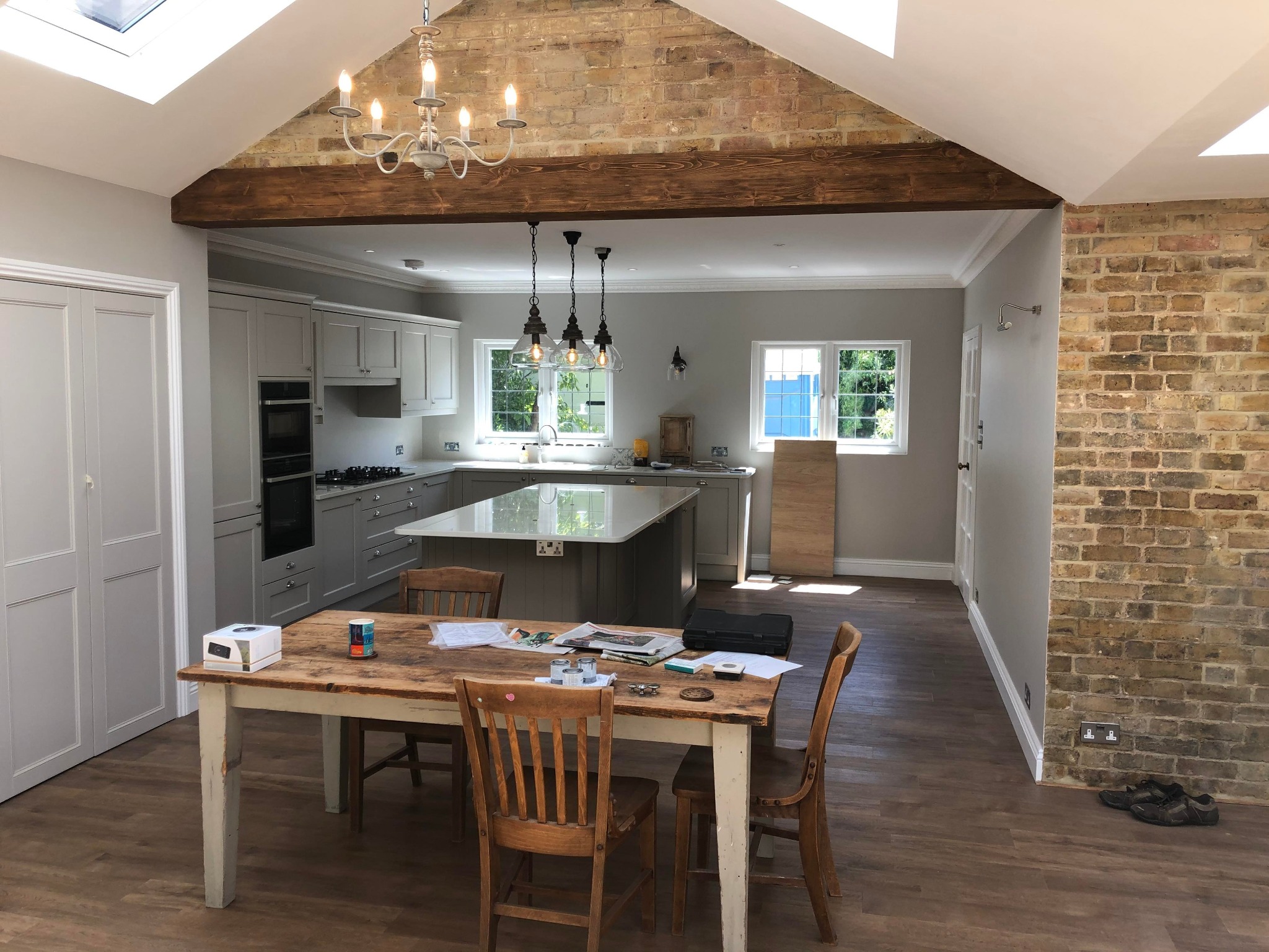 Jasnoszara kuchnia z wyspą i drewnianym stołem, ceglana ściana z drewnianą belką stropową, widoczne okna dachowe i wiszące lampy.
