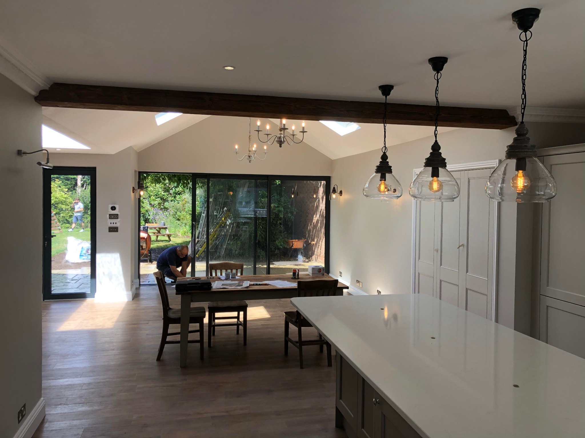 Przestronne wnętrze domu po remoncie z widoczną wyspą kuchenną, drewnianą podłogą, dużymi oknami przesuwnymi wychodzącymi na ogród i pracującym mężczyzną w tle, który coś montuje.