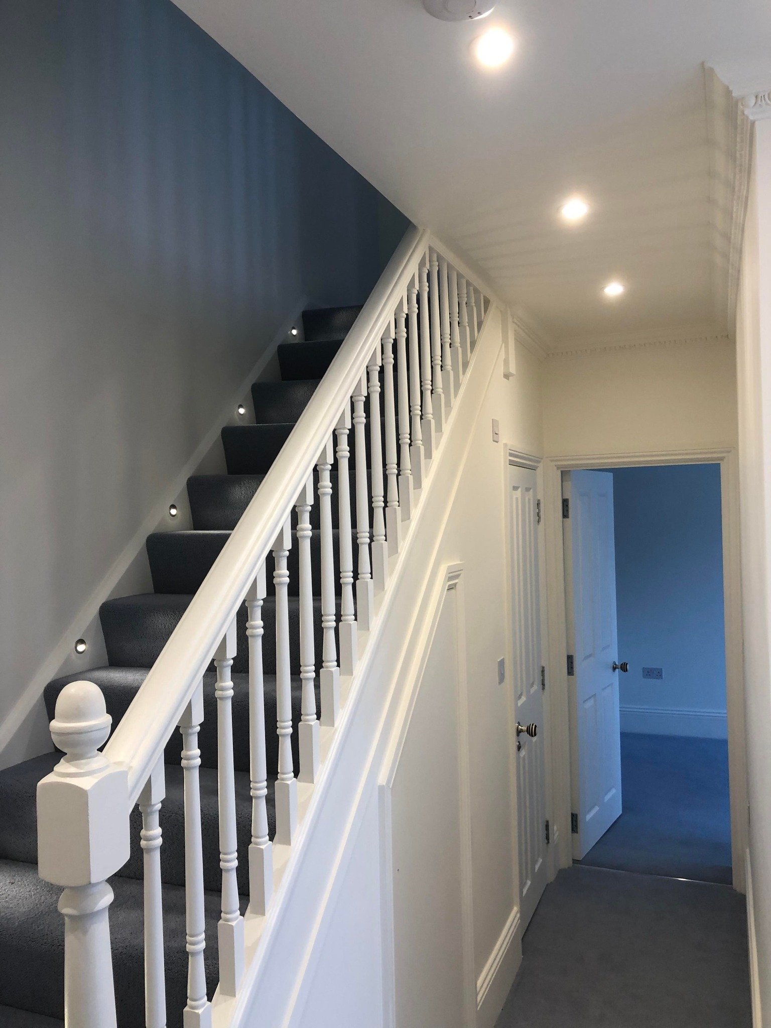 Klatka schodowa z szarymi schodami i białymi balustradami, oświetlona punktowymi lampkami w ścianie, z widokiem na otwarte drzwi do pokoju pomalowanego na niebiesko.