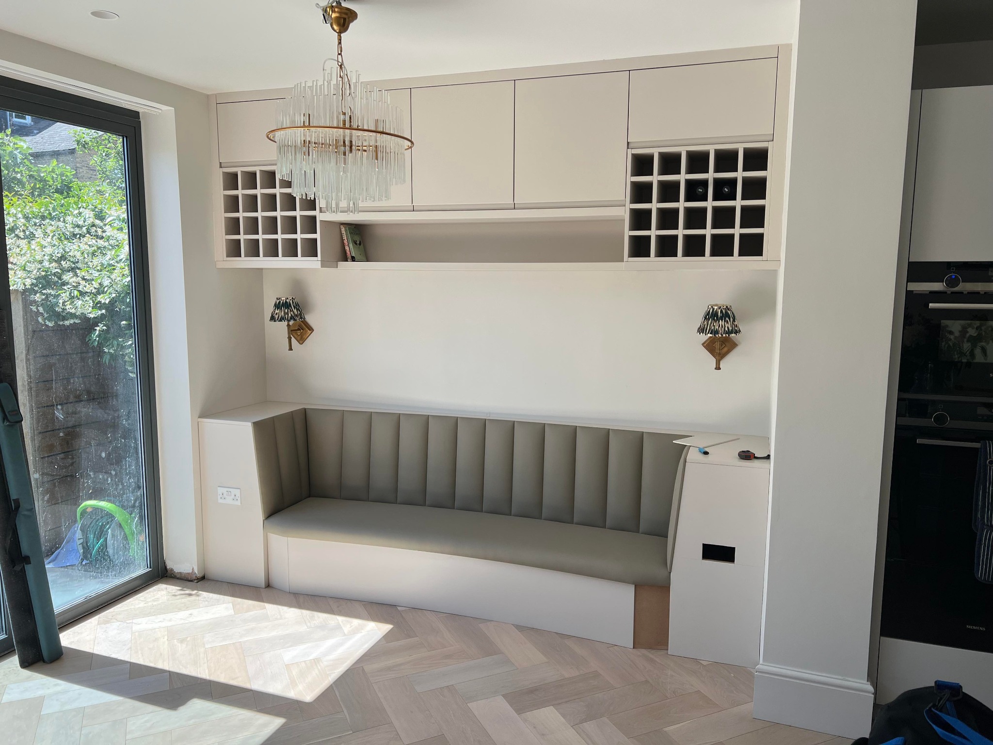 Wnęka z tapicerowanym siedziskiem i szafkami nad nią, podświetlona ozdobnym żyrandolem i kinkietami, na podłodze parkiet ułożony w jodełkę, widoczne ślady prac wykończeniowych.