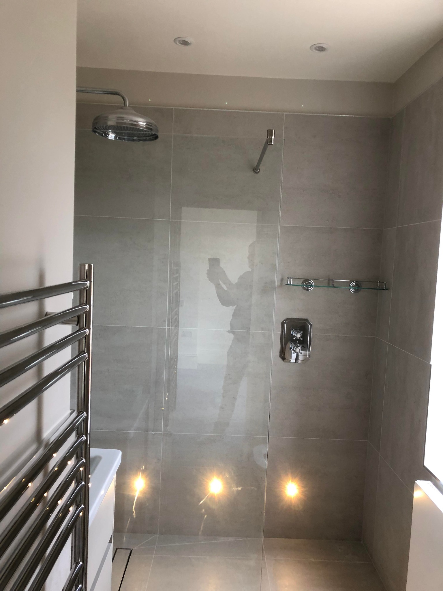 Nowoczesna kabina prysznicowa z deszczownicą, grzejnikiem drabinkowym i szklaną półką, wykończona szarymi płytkami ceramicznymi z odbijającym się w nich światłem.