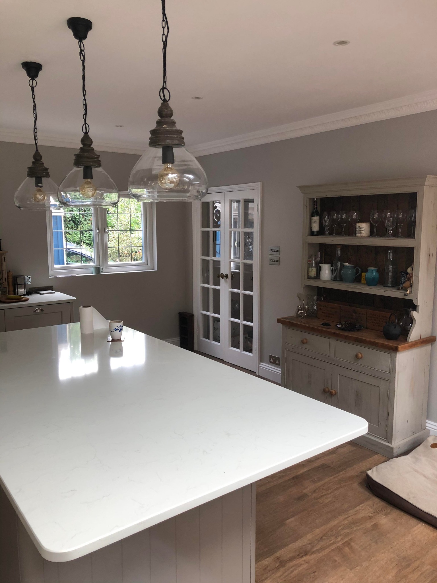 Jasna kuchnia z białym blatem wyspy, trzema wiszącymi lampami z przezroczystego szkła i drewnianymi elementami, oraz kredensem w stylu vintage z przeszklonymi drzwiami w tle.