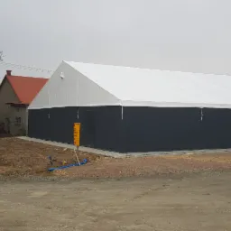 Zewnętrzny widok hali z płyt warstwowych o grafitowych ścianach i białym dachu, zlokalizowanej w otoczeniu wiejskim, z widocznymi przewodami instalacji na zewnątrz.