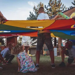 Dzieci bawią się pod kolorową chustą animacyjną trzymaną przez dorosłych na trawie podczas imprezy plenerowej, widoczne drzewa i budynek w tle.