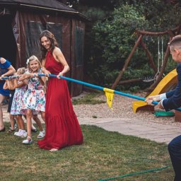 Animacje dla dzieci www.super-zabawa.pl - Grupa osób, w tym dzieci i dorośli, bierze udział w przeciąganiu liny na trawniku podczas imprezy plenerowej, z drewnianym domkiem i placem zabaw w tle.