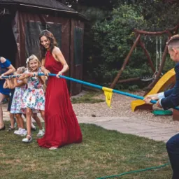 Grupa osób, w tym dzieci i dorośli, bierze udział w przeciąganiu liny na trawniku podczas imprezy plenerowej, z drewnianym domkiem i placem zabaw w tle.