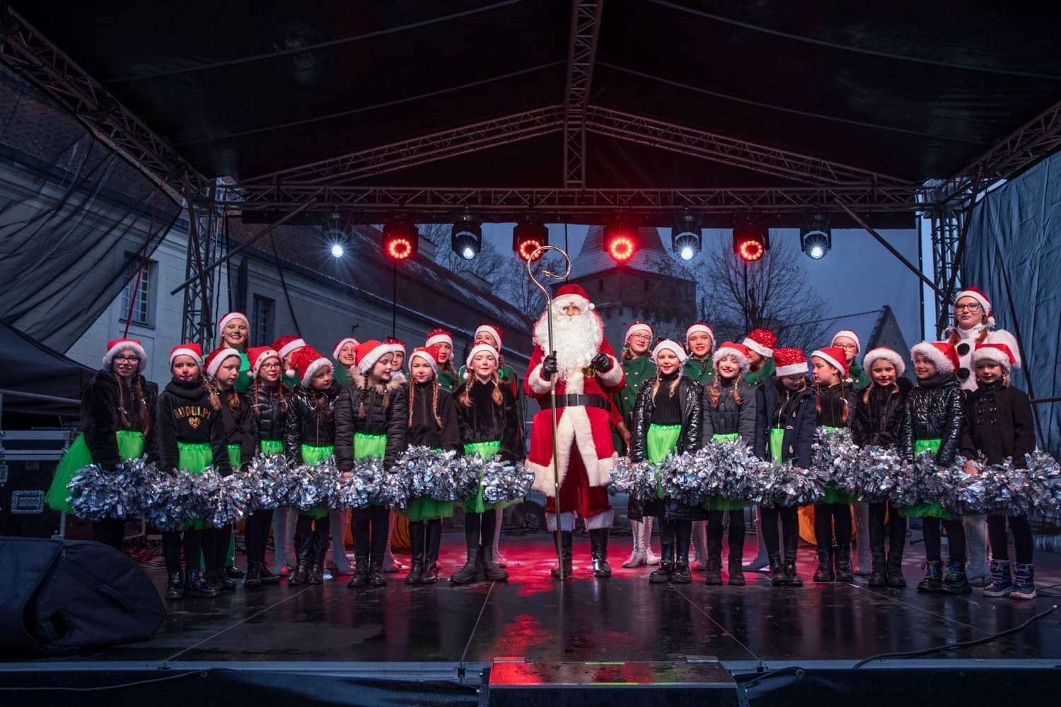 Występ grupy dzieci w strojach świątecznych z Mikołajem na scenie w Łaznowskiej Woli. Dzieci trzymają srebrne pompony, a w tle widać budynek.