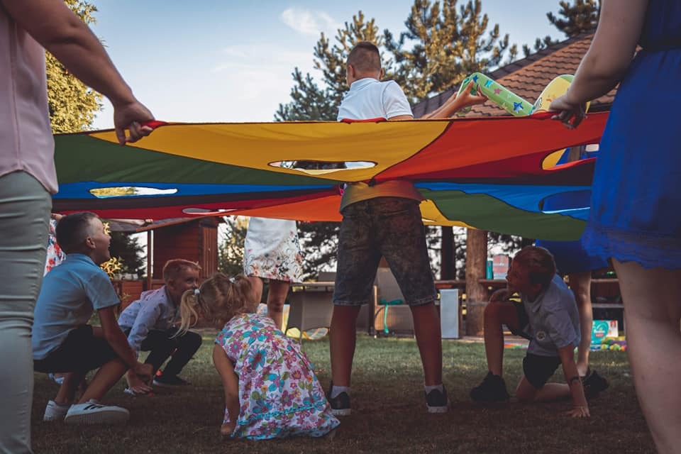 Dzieci bawią się pod kolorową chustą animacyjną trzymaną przez dorosłych na trawie podczas imprezy plenerowej, widoczne drzewa i budynek w tle.