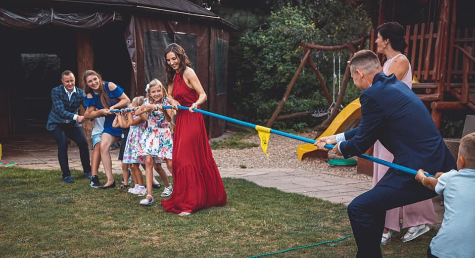 Grupa osób, w tym dzieci i dorośli, bierze udział w przeciąganiu liny na trawniku podczas imprezy plenerowej, z drewnianym domkiem i placem zabaw w tle.