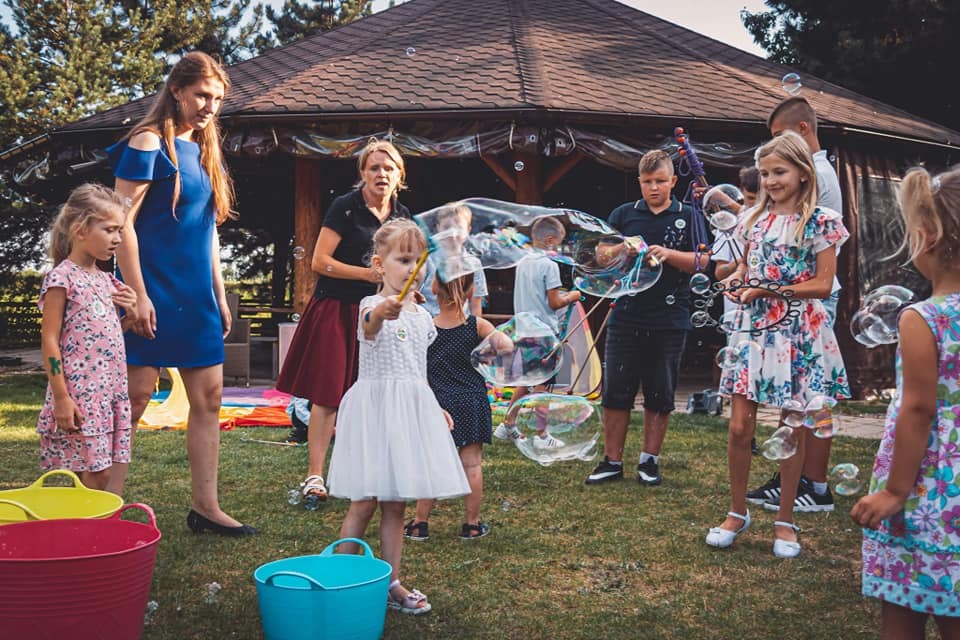 Grupa dzieci i dorosłych puszczających bańki mydlane na zewnątrz, w tle altana z brązowym dachem, kolorowe wiaderka na trawie.