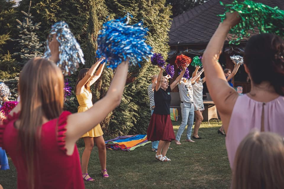 Grupa osób w różnym wieku, trzymających kolorowe pompony w górze, prawdopodobnie podczas animacji lub imprezy na świeżym powietrzu, z zielonym trawnikiem i domem w tle.