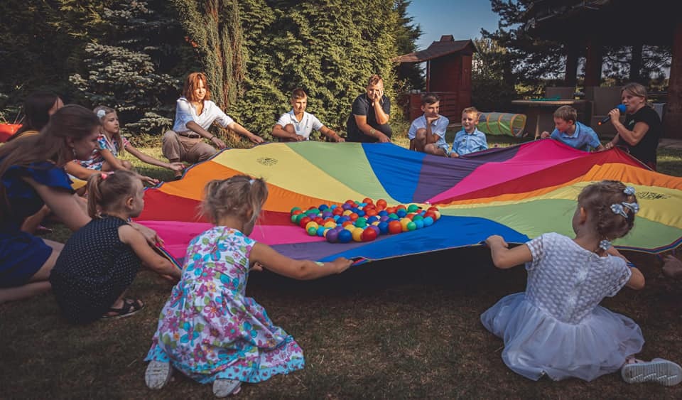 Grupa dzieci i dorosłych trzymająca kolorową chustę animacyjną z piłeczkami w środku podczas zabawy na trawie w ogrodzie, widoczna kobieta z mikrofonem, z tyłu dom i drzewa.