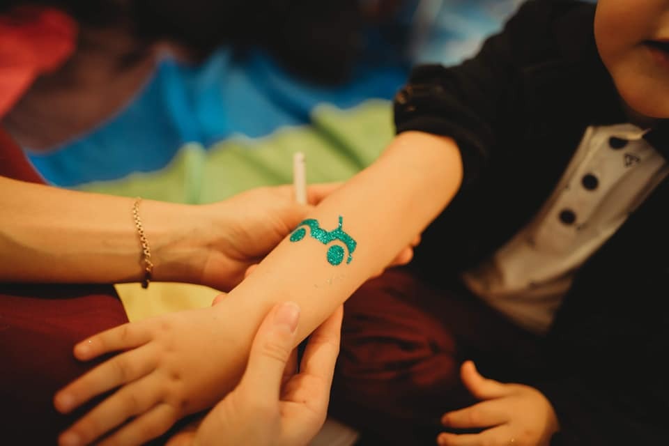 Błyszczący, turkusowy tatuaż brokatowy w kształcie traktora na ramieniu dziecka, aplikowany przez osobę dorosłą. Chłopiec ubrany w koszulę z motywem muchy.