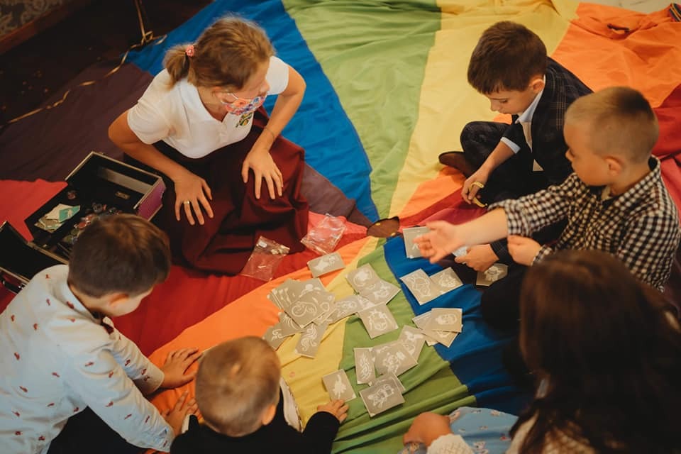 Grupa dzieci siedzi na tęczowej chuście, grając w karty z ilustracjami, obok leży otwarty kuferek z rekwizytami.