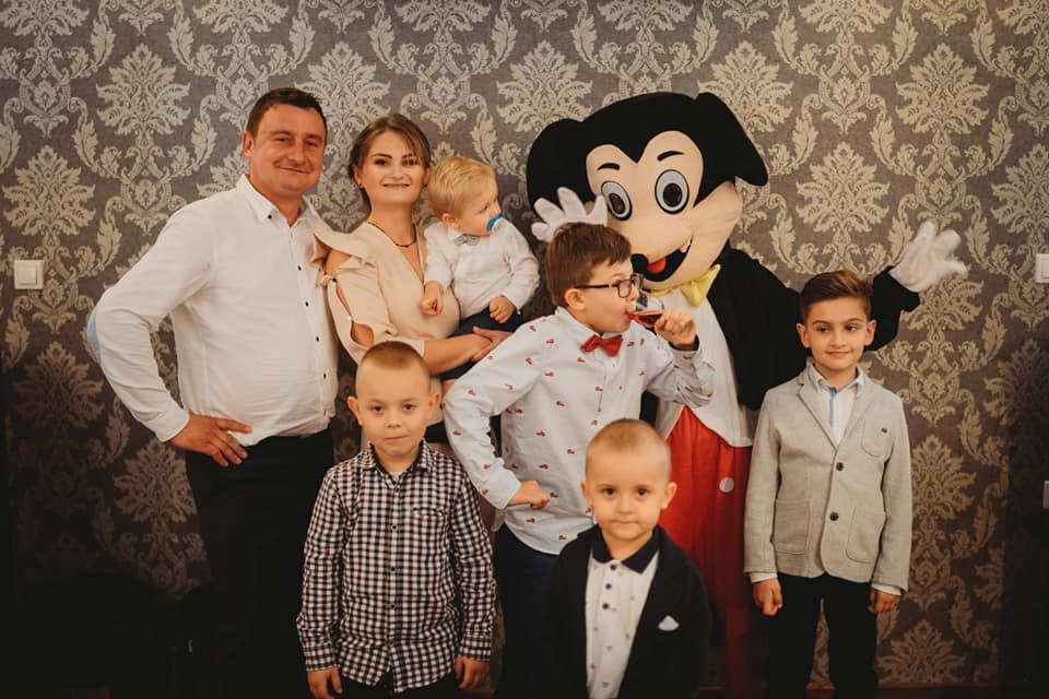 Grupa ludzi, w tym dzieci i osoba w stroju Myszki Miki, pozują do zdjęcia na tle wzorzystej tapety w pomieszczeniu.