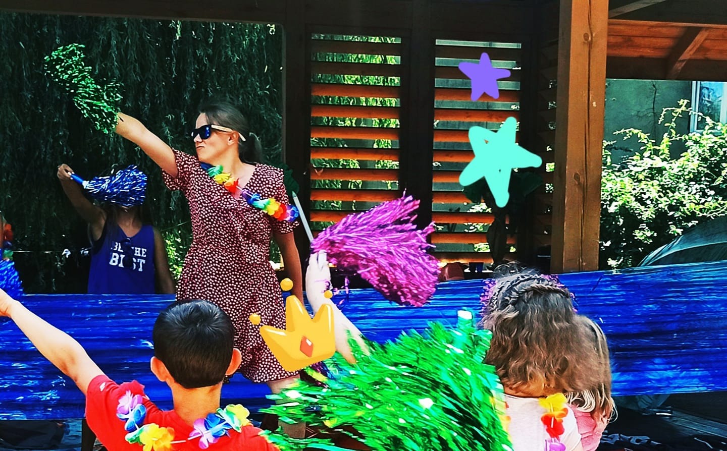 Kobieta w okularach i wzorzystej sukience, z pomponami w dłoniach, prowadzi animację dla dzieci podczas imprezy plenerowej, w tle widoczne dzieci i dekoracje.