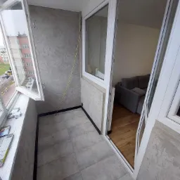 Wykończony balkon z szarymi płytkami podłogowymi, ścianami o fakturze betonu, otwartym oknem z widokiem na budynek mieszkalny i otwartymi drzwiami balkonowymi, przez które widać część salonu...