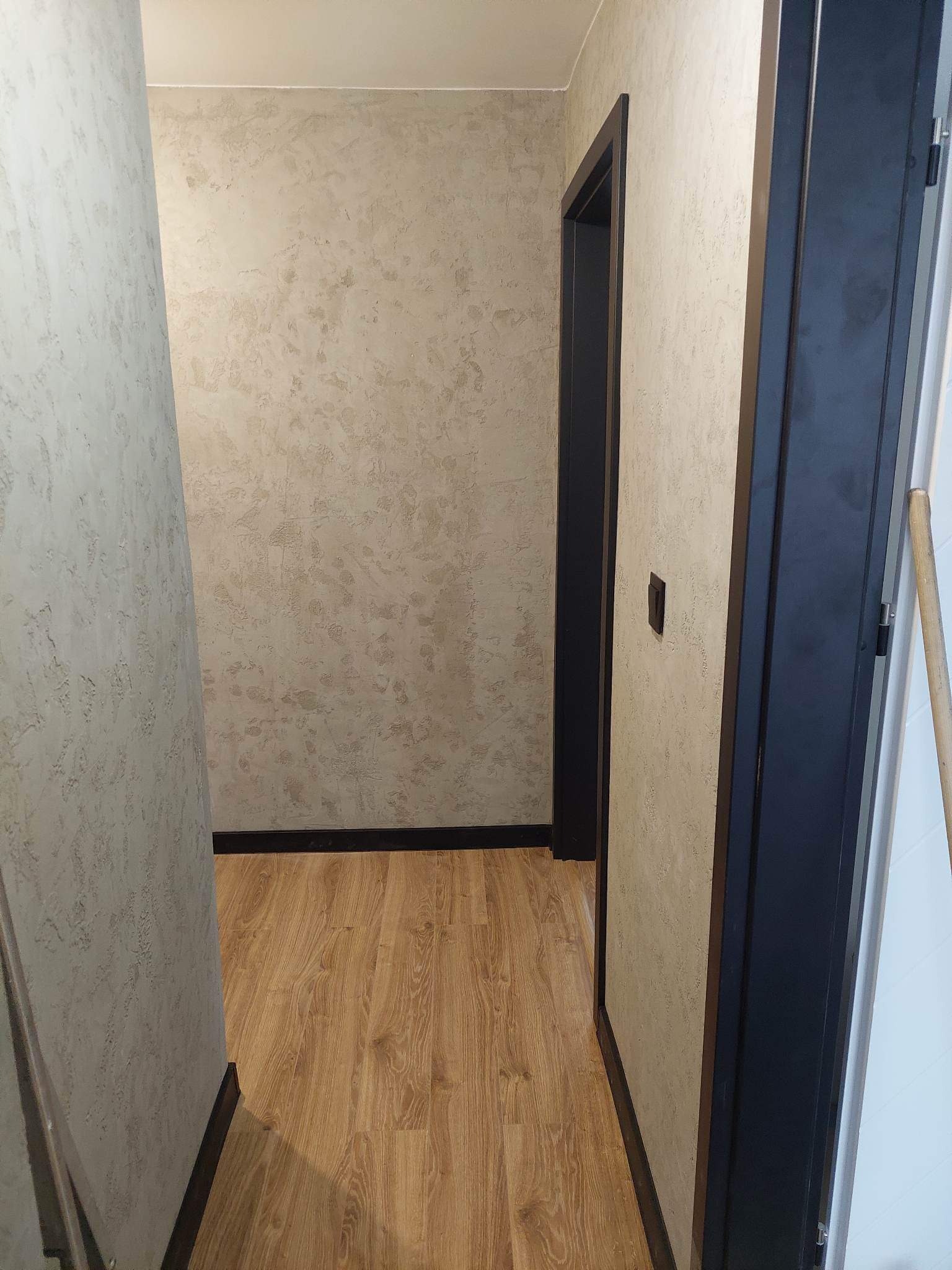 Korytarz po remoncie z beżową ścianą o nieregularnej fakturze, drewnopodobną podłogą i ciemnymi futrynami drzwi.