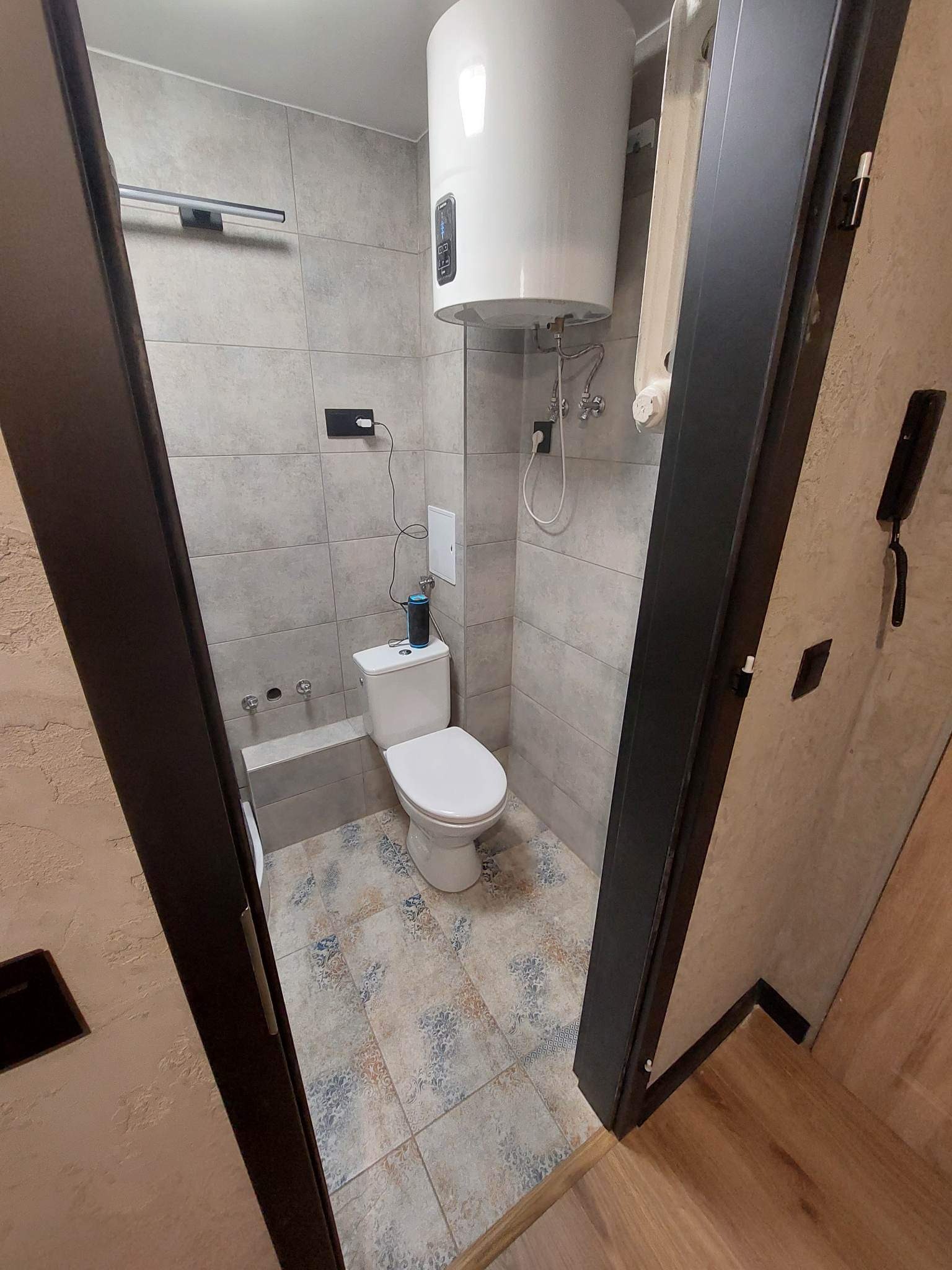 Nowoczesna łazienka z szarymi płytkami, wzorzystą podłogą, białym bojlerem i toaletą oraz czarnym inteligentnym głośnikiem na spłuczce.