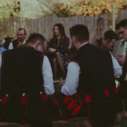 Zespół muzyczny w strojach ludowych grający na akordeonie i innych instrumentach podczas wydarzenia plenerowego, w tle siedząca kobieta i drewniany płot.