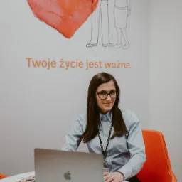 Kobieta w okularach siedzi przy biurku z laptopem, w tle rysunek rodziny i serce z napisem 'Twoje życie jest ważne'.