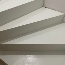 Woodstrug - Białe schody pokryte błyszczącą, jednolitą powłoką, widoczne z dołu pod kątem, z niedoskonałościami wykończenia przy ścianie.
